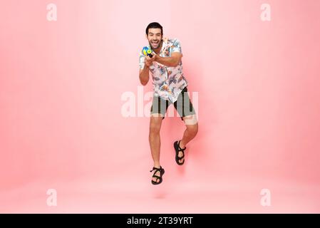 Bellissimo e sorridente turista caucasico che gioca con la pistola ad acqua e salta in uno sfondo rosa studio per il festival Songkran in Thailandia An Foto Stock