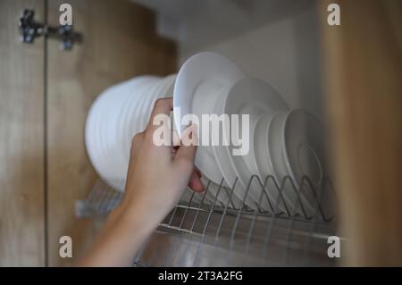 La mano della donna tira i piatti bianchi puliti fuori dall'armadietto della cucina. Concetto di servizi di aiuto per la casa Foto Stock