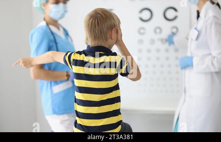 Ragazzo si siede con la schiena all'appuntamento dell'oftalmologo, guarda il tavolo e risponde alle domande. Diagnosi del concetto di visione nei bambini Foto Stock