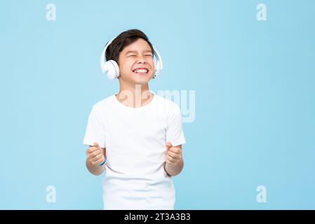 Sorridente, sorridente, ragazzo asiatico che indossa le cuffie, ascolta musica e pugni clandestini in uno studio di registrazione isolato di colore blu chiaro Foto Stock