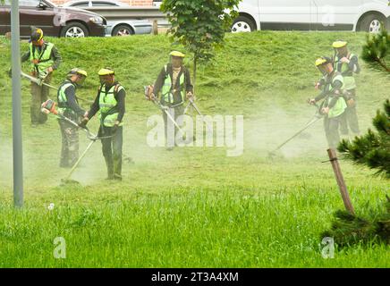 Ucraina, Kiev, 06.20.2019. Un gruppo di lavoratori municipali pubblici in uniformi verdi e maschere falciando erba in un parco con Handheld Gasoline Lawn mo Foto Stock