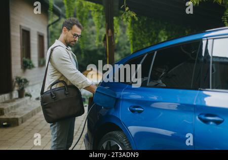 Ritratto di un uomo d'affari che carica la sua auto elettrica davanti a casa. Collegare il caricabatteria alla porta di carica. Foto Stock