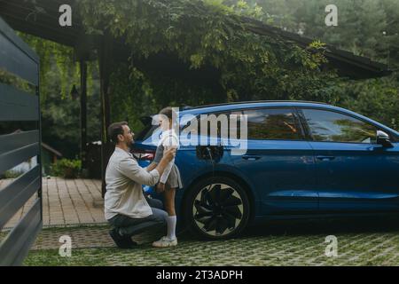 Ritratto di padre e figlia che parlano, mentre si carica un'auto elettrica di famiglia di fronte a casa. Veicolo elettrico con caricabatterie nella porta di carica. Foto Stock