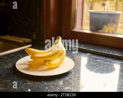 Banane fresche su un piatto bianco sopra il bancone della cucina in granito nero. Foto Stock