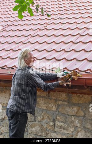 Un uomo maturo rimuove foglie e detriti dalla fossa della sua casa. Pulizia di uno scarico della pioggia. Foto Stock