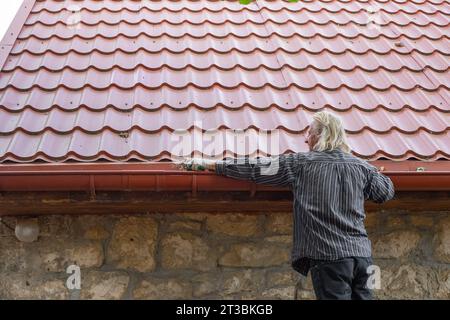 Un uomo maturo rimuove foglie e detriti dalla fossa della sua casa. Pulizia di uno scarico della pioggia. Foto Stock