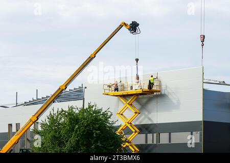 Montaggio a parete con pannelli sandwich utilizzando una gru telescopica a braccio e una piattaforma di sollevamento a forbice semovente gialla Foto Stock