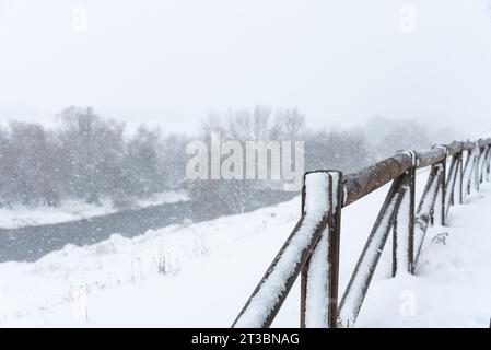 Recinzione in legno coperta dalla neve accanto a un fiume sotto una forte nevicata in inverno Foto Stock