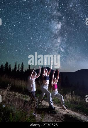 Tre ragazze che fanno yoga la notte stellata. Ragazze sportive che fanno ginnastica con vista sulla via Lattea. Esercizi di yoga in montagna. Foto Stock