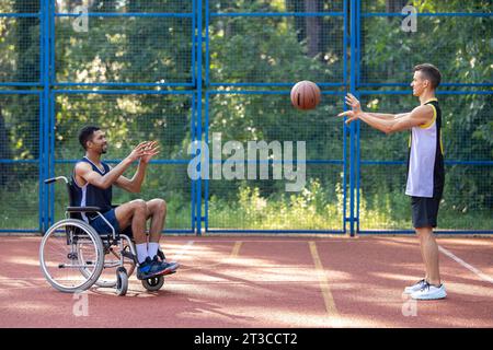 Uomo in sedia a rotelle e suo amico che si diverte giocando a basket all'aperto. Foto Stock