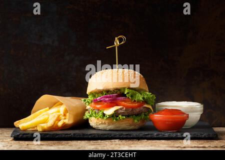 Hamburger con cotoletta di manzo, patatine fritte, salse su un piatto di pietra nera su sfondo scuro. Fast food, hamburger Foto Stock