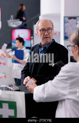 Un vecchio che chiede alla cassa della farmacia di scansionare il prezzo della crema solare alla reception. Dipendente farmaceutico che fornisce assistenza al cliente e utilizza lo scanner per codici a barre della crema solare Foto Stock