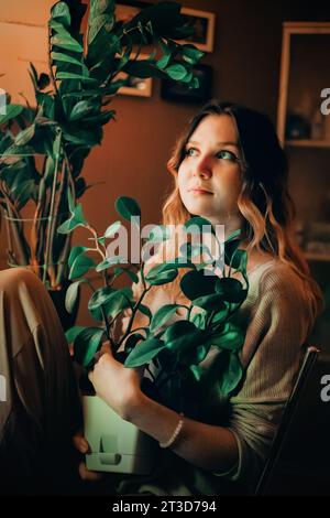 Una giovane donna in abbigliamento informale seduta a casa con un vaso tra le mani, circondata da altre piante. Mette in mostra i temi dell'ecologia e della carina Foto Stock