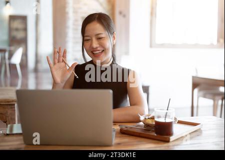 Una bella e allegra donna asiatica sta partecipando a un incontro online con il suo team mentre lavora in remoto al bar, salutando la sua mano per salutarlo Foto Stock