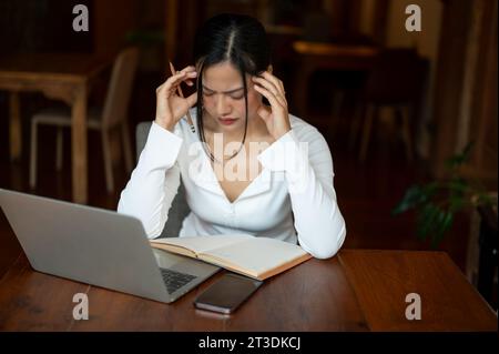 Una donna asiatica stressata che lavora a distanza in un bar, soffre di mal di testa mentre legge un libro, lavora su un progetto difficile. Foto Stock