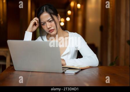 Una donna asiatica millenaria, stressata e seria, sta guardando lo schermo del suo notebook con un volto insoddisfatto, avendo un problema con il suo progetto mentre lavora Foto Stock