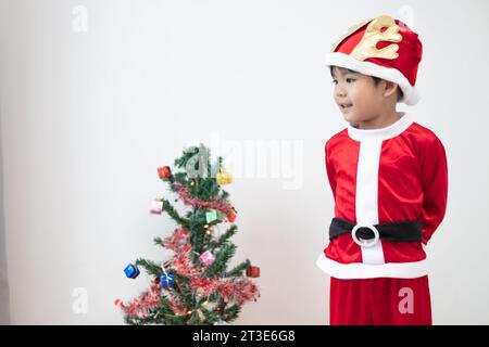 Ragazzo asiatico che indossa il costume di Babbo Natale in piedi e gioca vicino a Un divertente albero di Natale su sfondo bianco. Foto Stock