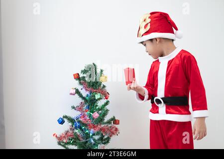 Ragazzo asiatico che indossa il costume di Babbo Natale in piedi e gioca vicino a Un divertente albero di Natale su sfondo bianco. Foto Stock