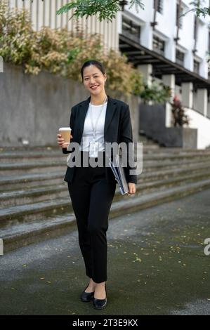 Un ritratto di una donna d'affari asiatica millenaria sicura di sé e attraente in un abito da lavoro formale con una tazza da caffè da asporto e leganti nelle sue mani Foto Stock