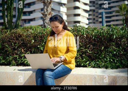 Giovane donna adulta multietnica vestita con abiti informali, pianificando una Start-up, digitando testo sul laptop mentre si lavora in telelavoro, lavorando online all'aperto. Persone. Foto Stock