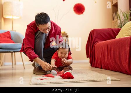 Ritratto a lunghezza intera di un padre asiatico felice che gioca con un grazioso bambino seduto sul pavimento a casa e appeso decorazioni di carta rossa per la celebrazione del capodanno cinese, spazio fotocopie Foto Stock