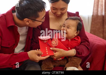 Ritratto della felice famiglia asiatica con un bambino carino che si gode la celebrazione del Capodanno cinese a casa e che tiene le buste rosse hanno abbondanza traboccante ogni anno Foto Stock