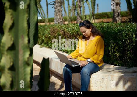 Giovane afroamericana bella donna in abiti casual, programmatore freelance che lavora su una nuova startup, usando un computer portatile, seduto su una panchina di pietra in una natura Foto Stock