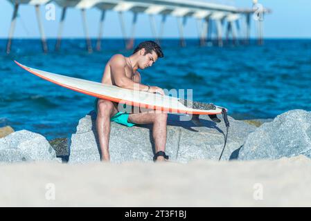 Un giovane serio che pulisce la tavola da surf sulla spiaggia soleggiata Foto Stock