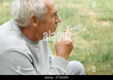 Uomo anziano che accende una sigaretta all'aperto su sfondo verde. Vecchio uomo premuroso che fuma, guardando da parte. Faccia da vicino. Pensione rilassata Foto Stock