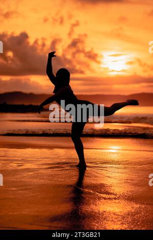 Silhouette di una donna asiatica che danza balletto con grande flessibilità e vista delle onde dietro di lei al tramonto Foto Stock