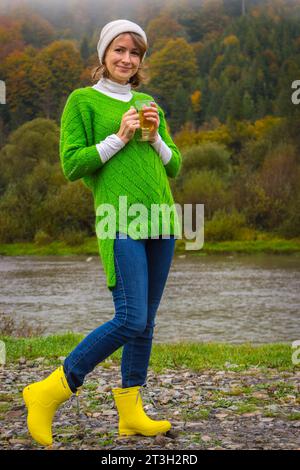 Donna sulle montagne autunnali sulla riva del fiume. Ragazza in abiti caldi sulla riva del fiume con una tazza di tè caldo. Viaggia tra le montagne dei Carpazi, Ucraina. Foto Stock