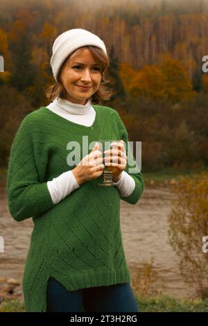 Donna sulle montagne autunnali sulla riva del fiume. Ragazza in abiti caldi sulla riva del fiume con una tazza di tè caldo. Viaggia tra le montagne dei Carpazi, Ucraina. Foto Stock