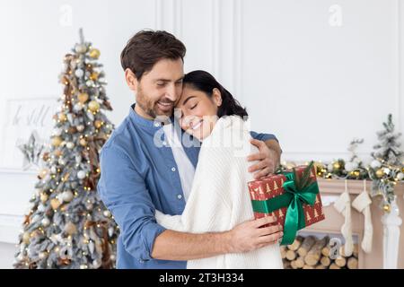 Una giovane coppia, una famiglia, un uomo e una donna festeggiano insieme Natale e le vacanze invernali a casa, abbracciano l'albero di Natale, scambiano regali, si congratulano l'un l'altro. Foto Stock