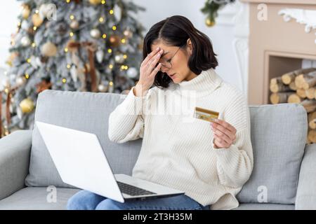 Triste giovane donna seduta a casa sul divano per le vacanze vicino all'albero di Natale, con una carta di credito e con la testa in preda alla preoccupazione. Foto Stock