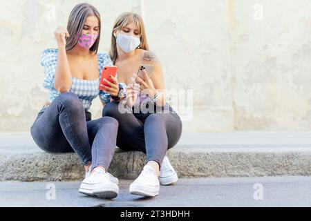 donne serie con una maschera che guardano uno smartphone, controllano le notizie su un marciapiede Foto Stock