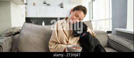 donna con i capelli corti seduta su un divano a casa, che mangia cereali a colazione e accarezza il suo amato cane. Il cane sta annusando una ciotola di cereali Foto Stock