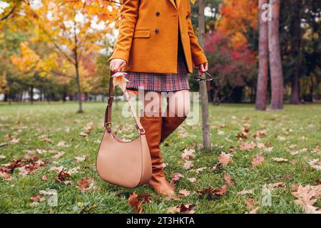 Stivali alti al ginocchio arancioni. Donna alla moda che indossa un blazer  elegante minigonna a quadri che cammina nel parco autunnale con una borsa  beige tra le foglie. Copia spazio Foto stock 