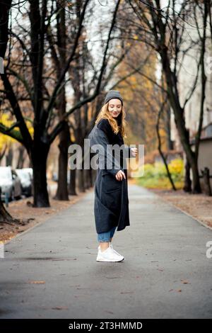 Nel bel mezzo di un paesaggio urbano autunnale adornato da foglie gialle, una giovane donna vestita di un lungo cappotto grigio e con uno zaino sorride brillantemente, embodyi Foto Stock