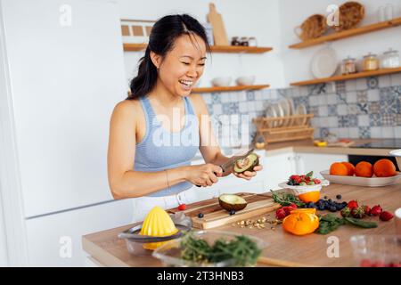 Una bella donna asiatica sorride e cucina la colazione in cucina. Una giovane affascinante donna coreana ride e taglia verdure e pesce. Sana f Foto Stock