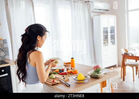 Una bella donna asiatica sorride e cucina la colazione in cucina. Una giovane affascinante donna coreana ride e taglia verdure e pesce. Sana f Foto Stock