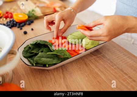 Una bella donna asiatica sorride e cucina la colazione in cucina. Una giovane affascinante donna coreana ride e taglia verdure e pesce. Sana f Foto Stock