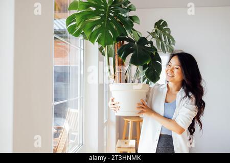 Un'attraente donna asiatica in abiti casual tiene una pentola con una pianta domestica. Un'affascinante giovane donna coreana si prende cura di un fiore monstera nei vivi Foto Stock