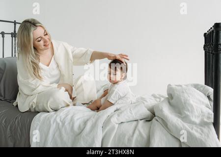 Ritratto di una bambina seduta accanto a sua madre sul letto la mattina dopo il sonno. Mamma raddrizza una ciocca di capelli per sua figlia. Co. Famiglia Foto Stock