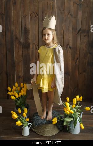 Ritratto di una principessa ragazza in una corona di cartone con una spada in mano, accanto a mazzi di tulipani gialli. Telaio verticale. Guarda la telecamera. Foto Stock