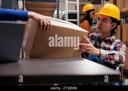 Commissionatore Storehouse su sedia a rotelle per l'evasione degli ordini e il prelievo di scatole di cartone dalle mani dei colleghi. Uomo asiatico con disabilità che gestisce le operazioni di servizio postale in un magazzino industriale Foto Stock