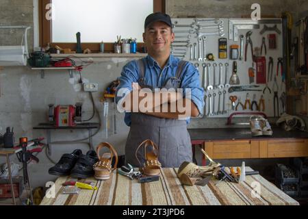 Immagine di un calzolaio artigiano sorridente che posa nel suo laboratorio mentre ripara le scarpe. Foto Stock