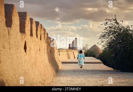 Giovane donna in abito etnico con ornamento blu che cammina alle mura della città di Itchan Kala, antica città di Khiva in Uzbekistan. Foto Stock
