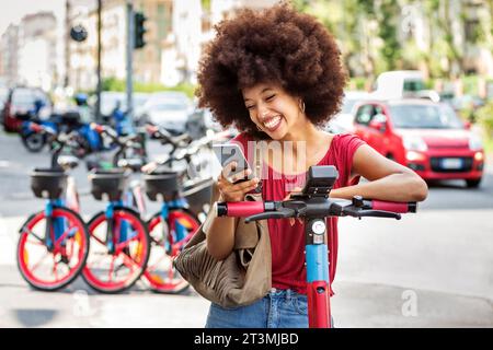 Felice donna nera con acconciatura afro che noleggia uno scooter elettrico in città con l'app sullo smartphone durante il giorno Foto Stock