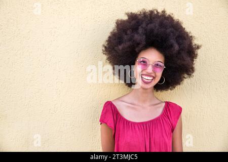 Ritratto di una giovane donna marocchina felice con acconciature afro ed eleganti occhiali da sole in piedi vicino alle pareti beige e guardando la macchina fotografica Foto Stock