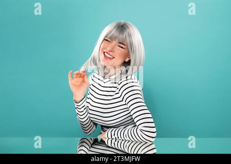 Giovane modello femminile positivo con camicia a maniche lunghe a righe che tocca i capelli grigi e gli occhi di chiusura mentre si siede al tavolo a specchio in studio Foto Stock